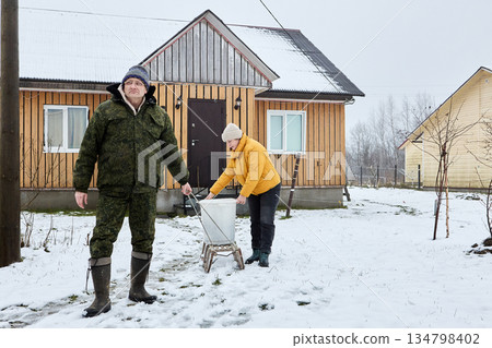 Man pulls sled by rope leaving rural house, woman holds garbage bin standing on it. 134798402