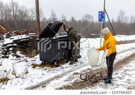 Collecting waste in snowy village, man lifts dumpster lid while companion waits with bucket on sled near muddy rural road. 134798407