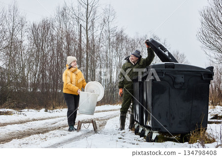 Large garbage dumpsters on wheels at waste collection point in village, mature couple delivered garbage bucket on sled. Large garbage dumpsters on wheels at waste collection point in village, mature couple delivered garbage bucket on sled. 134798408