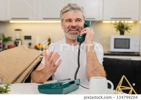 Middle-aged man answers retro landline phone smiling nostalgia hello kind call at sofa table at home 134799204