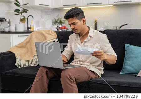 Indian man updates bill numbers in laptop spreadsheet checks docs and confirms monthly budget total Indian man updates bill numbers in laptop spreadsheet checks docs and confirms monthly budget total 134799229