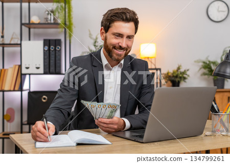 Middle-aged businessman counts cash dollars records budget on laptop writes notes in notebook plans Middle-aged businessman counts cash dollars records budget on laptop writes notes in notebook plans 134799261