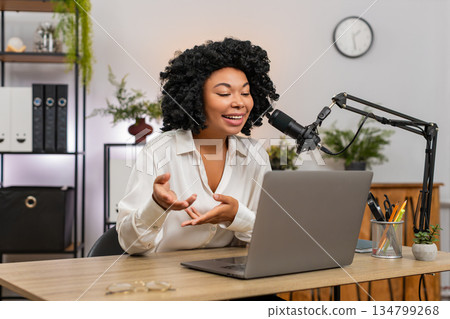 African American businesswoman gestures speaking into studio microphone recording podcast at laptop 134799268
