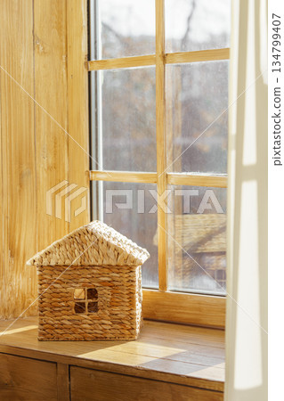 Small woven house model on sunny wooden window sill against natural light window in child room 134799407