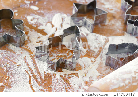 Baking gingerbread cookie cutters, wooden rolling pin, and flour are messily spread on kitchen table 134799418