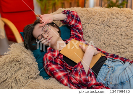 Young woman falling asleep with book in hand at home, peaceful expression and tired body language 134799480