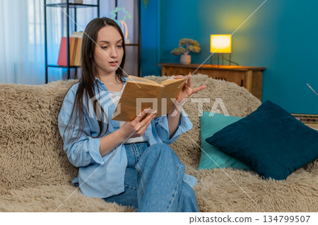 Young excited woman reading book on sofa deeply immersed emotionally reacting changing expressions Young excited woman reading book on sofa deeply immersed emotionally reacting changing expressions 134799507