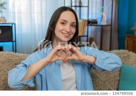 Cheerful young woman on sofa showing heart gesture happily expressing love friendship positivity Cheerful young woman on sofa showing heart gesture happily expressing love friendship positivity 134799508