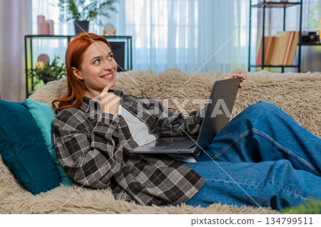 Woman sitting on sofa shopping online with laptop choosing dress touching chin in thoughtful pose Woman sitting on sofa shopping online with laptop choosing dress touching chin in thoughtful pose 134799511