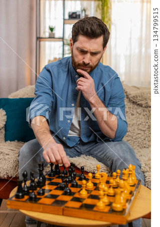 Young man playing chess alone making serious move deep thinking strategy concentration home sofa 134799515
