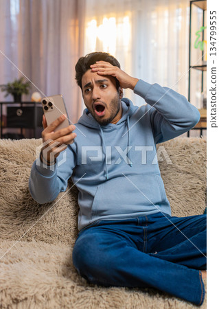 Shocked Indian young man reading bad news on smartphone phone while sitting on sofa in living room 134799555