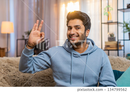 Happy Indian man showing OK gesture and smiling confidently while sitting on sofa at home room 134799562