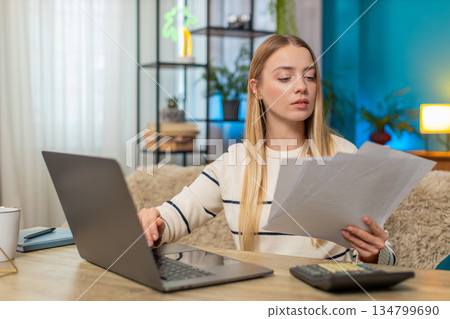 Caucasian woman managing tax declaration at home on sofa table, holding documents, calculator laptop 134799690
