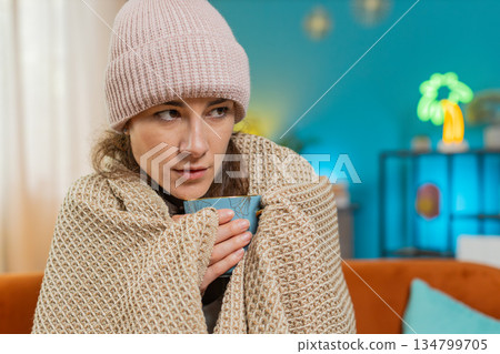 Young unhealthy sick ill woman covered by warm blanket drinks coffee freezes from cold flu at home Young unhealthy sick ill woman covered by warm blanket drinks coffee freezes from cold flu at home 134799705