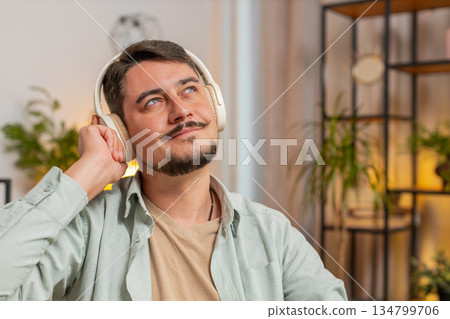 Happy relaxed overjoyed young Caucasian man in wireless headphones listening to music in home room Happy relaxed overjoyed young Caucasian man in wireless headphones listening to music in home room 134799706