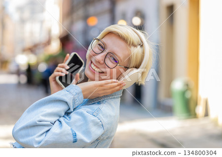 Carefree young woman in glasses listening music via wireless headphones while walking on city street Carefree young woman in glasses listening music via wireless headphones while walking on city street 134800304