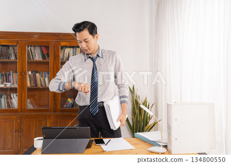 Male employee scrutinizing time as he handles paperwork at desk Male employee scrutinizing time as he handles paperwork at desk 134800550