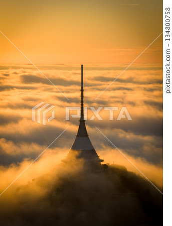 Aerial view of Jested Tower and Hotel rising above a sea of clouds at sunrise. Warm golden light and morning fog create a surreal, peaceful atmosphere over the Jizera Mountains in the Czech Republic. Aerial view of Jested Tower and Hotel rising above a sea of clouds at sunrise. Warm golden light and morning fog create a surreal, peaceful atmosphere over the Jizera Mountains in the Czech Republic. 134800758