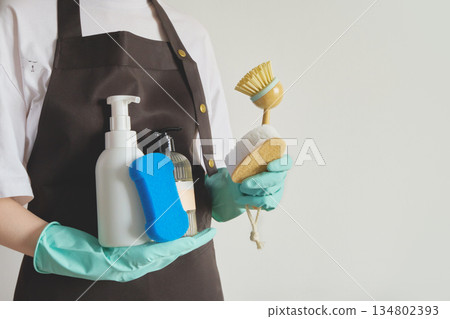 Person wearing an apron and gloves holding bottles, sponge and brush for cleaning Person wearing an apron and gloves holding bottles, sponge and brush for cleaning 134802393