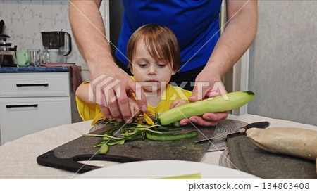 Father teaching toddler to peel cucumber in kitchen. Adult hands guiding a small child in peeling fresh cucumber with peeler at kitchen table, family bonding and learning concept. 134803408