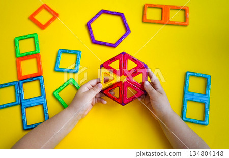 A child's creative play session with magnetic building blocks is captured against a bright yellow backdrop, showing various constructed shapes. 134804148