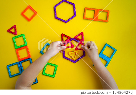 The photo shows a little girl creatively playing with magnetic building blocks, constructing various designs against a solid yellow background for contrast. 134804273
