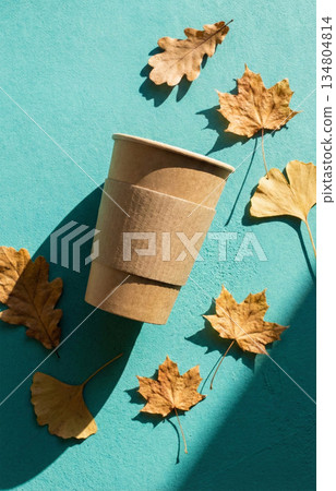 Eco coffee cup and autumn leaf against a teal background. Autumn paper coffee cup and fall leaves on vibrant teal background. 134804814