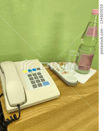 A bedside table in a room or hotel with a telephone, remote control, and a bottle of water A bedside table in a room or hotel with a telephone, remote control, and a bottle of water 134805050