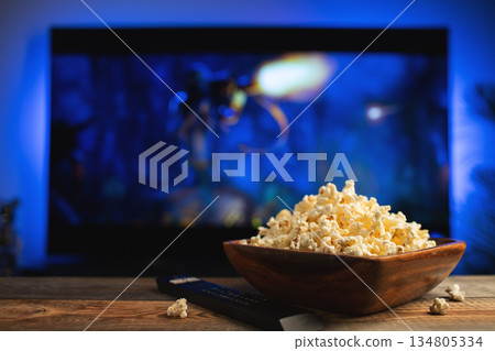 A wooden bowl of popcorn and remote control in the background the TV works. Evening cozy watching a movie or TV series at home A wooden bowl of popcorn and remote control in the background the TV works. Evening cozy watching a movie or TV series at home 134805334