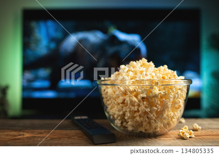 A glass bowl of popcorn and remote control in the background the TV works. Evening cozy watching a movie or TV series at home 134805335
