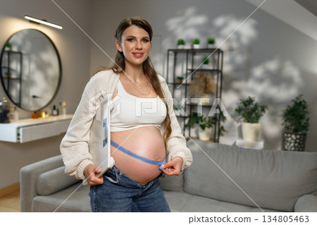 Pregnant woman stands in living room holding scale and measuring tape around her belly, monitoring weight gain during pregnancy 134805463