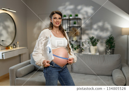 Smiling pregnant woman in casual clothes measures her growing belly with a tape measure in a cozy living room setting 134805526
