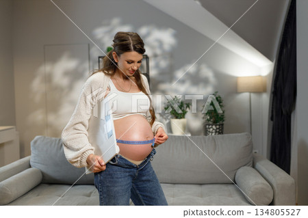 Pregnant woman measures her growing belly with a tape measure while holding a scale in a cozy living room setting 134805527