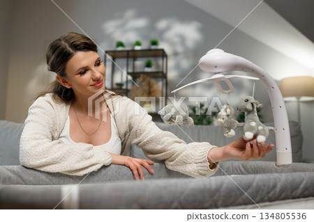 Young mother smiles gently while observing plush animal toys on a baby crib mobile in a softly lit nursery room Young mother smiles gently while observing plush animal toys on a baby crib mobile in a softly lit nursery room 134805536