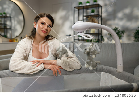 Young mother gazes thoughtfully near baby crib with mobile, creating serene nursery atmosphere 134805539