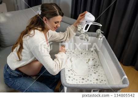 Expecting mother arranges nursery mobile above crib with baby clothes inside, anticipating newborn arrival Expecting mother arranges nursery mobile above crib with baby clothes inside, anticipating newborn arrival 134805542