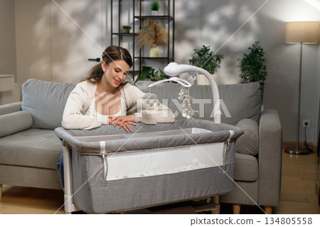 Young mother gazes lovingly at a crib with a mobile, creating a serene nursery atmosphere in a modern living room Young mother gazes lovingly at a crib with a mobile, creating a serene nursery atmosphere in a modern living room 134805558
