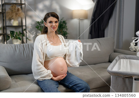 Expecting mother sits on couch holding tiny white onesie, anticipating newborn arrival with joy and tenderness in cozy home setting Expecting mother sits on couch holding tiny white onesie, anticipating newborn arrival with joy and tenderness in cozy home setting 134805560