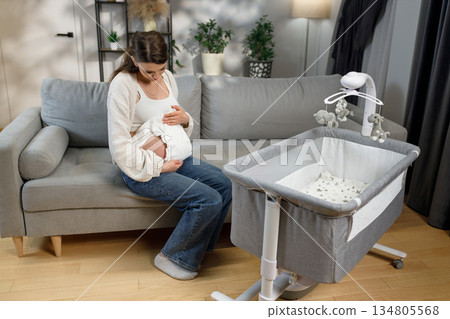 Pregnant woman sits on sofa holding belly near bedside bassinet with mobile, preparing for new baby arrival Pregnant woman sits on sofa holding belly near bedside bassinet with mobile, preparing for new baby arrival 134805568