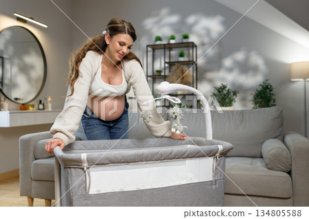 Expecting mother prepares nursery furniture, touching mobile above baby bassinet with gentle smile Expecting mother prepares nursery furniture, touching mobile above baby bassinet with gentle smile 134805588