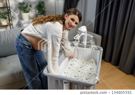 Smiling pregnant woman adjusts bedding in portable crib, preparing nursery for newborn baby arrival Smiling pregnant woman adjusts bedding in portable crib, preparing nursery for newborn baby arrival 134805591