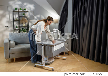 Pregnant woman arranging bedding in a bedside bassinet next to sofa in living room with plants and curtains Pregnant woman arranging bedding in a bedside bassinet next to sofa in living room with plants and curtains 134805595