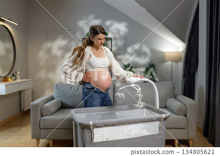 Expecting mother in casual clothes stands near crib, touching her belly and adjusting mobile, anticipating baby's arrival 134805621