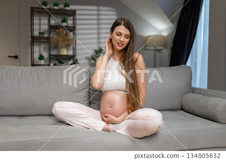 Young expectant mother sits cross-legged on a comfortable couch, cradling her belly with a gentle smile 134805632