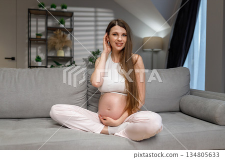 Young expectant mother sits comfortably on a couch, cradling her belly with a gentle smile, anticipating new life 134805633