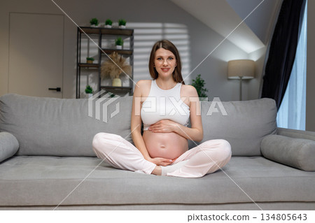 Young expectant mother sits cross-legged on a comfortable sofa, cradling her pregnant belly with a gentle smile 134805643