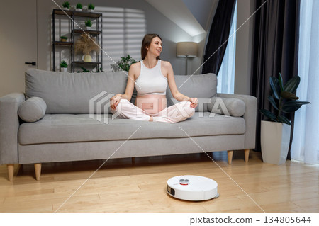 Pregnant woman sits cross-legged on sofa while robot vacuum cleans floor in modern living room, creating peaceful atmosphere 134805644