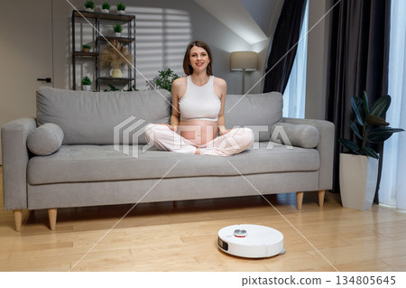 Pregnant woman sits cross-legged on sofa while robot vacuum cleans floor, effortless home maintenance 134805645