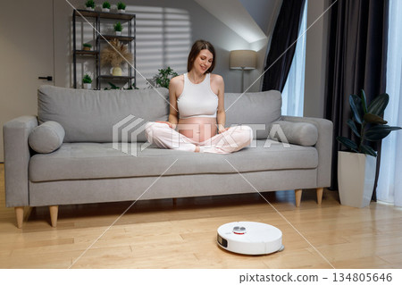 Pregnant woman sits cross-legged on sofa while robot vacuum cleans floor in modern living room 134805646