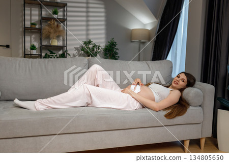 Expecting mother rests comfortably on a plush couch, cradling her belly with a gentle smile Expecting mother rests comfortably on a plush couch, cradling her belly with a gentle smile 134805650